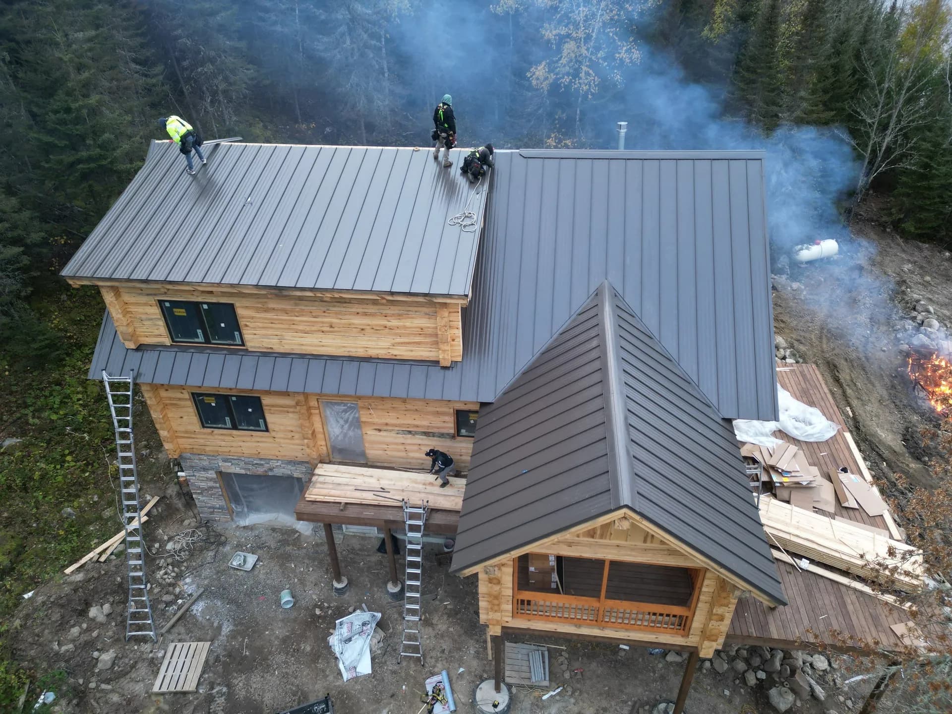 Aerial view of a large timber-frame lodge near Crane Lake with new charcoal-gray standing seam metal roof, crew installing panels, wood smoke rising from a brush pile in the background, October 2023