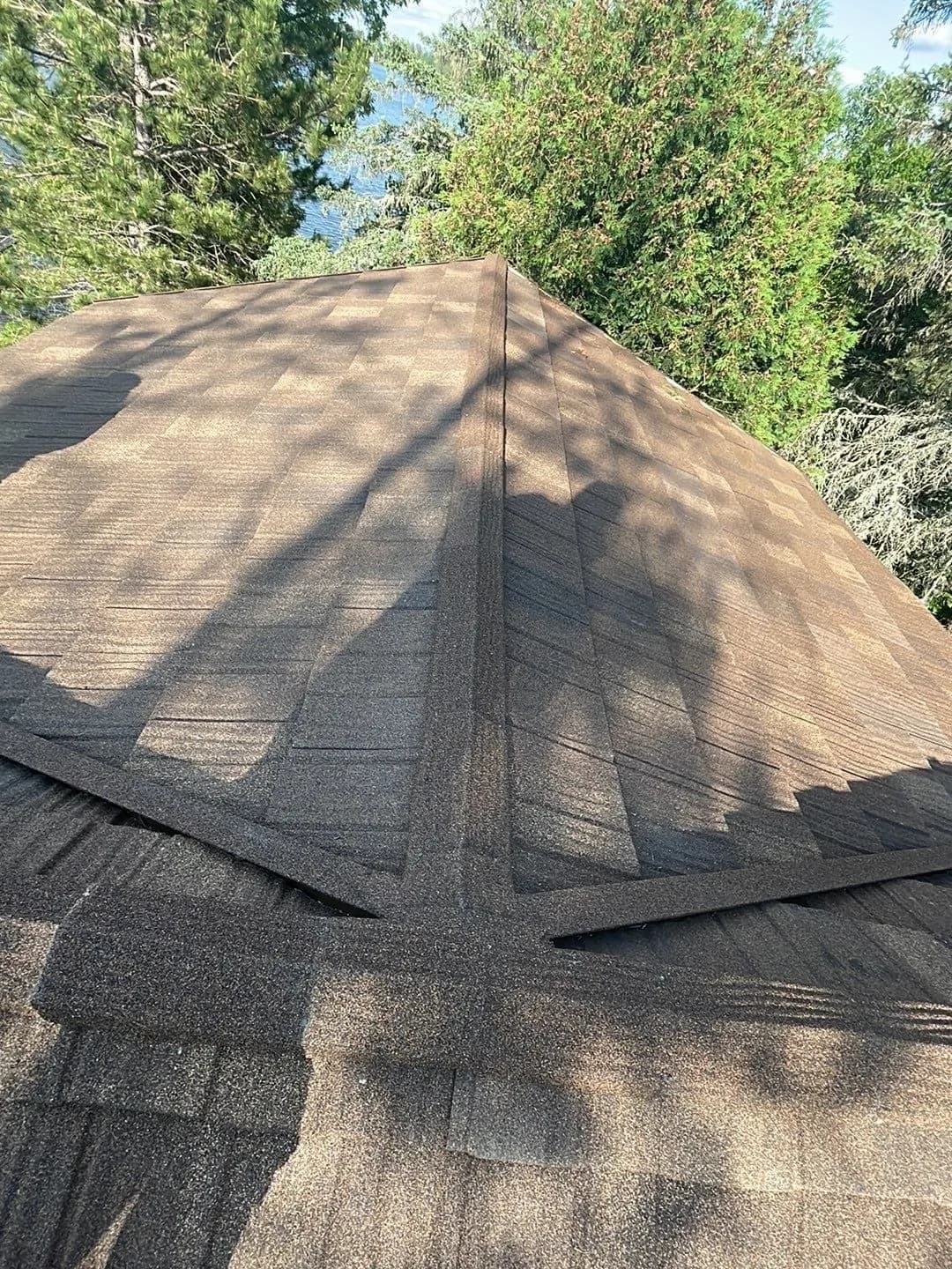 Dark architectural asphalt shingle roof on a lake cabin garage gable in northern Minnesota, satellite dish mount, blue sky with light clouds, birch and pine trees, June 2023